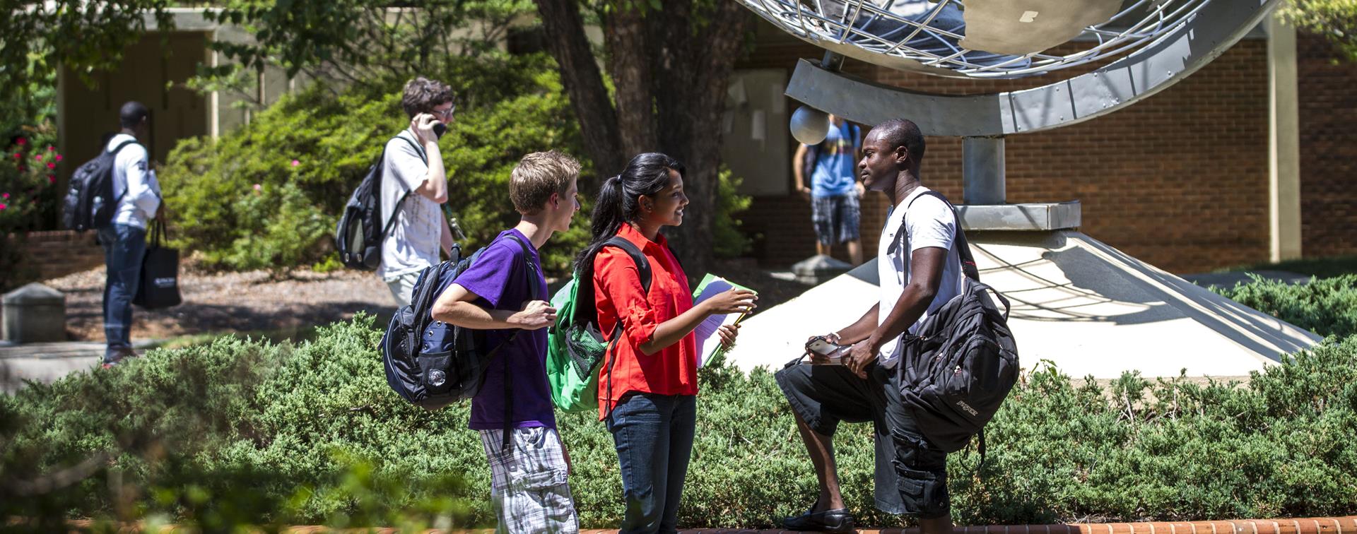 Picture of three students on campus having a conversation in front of a globe.
