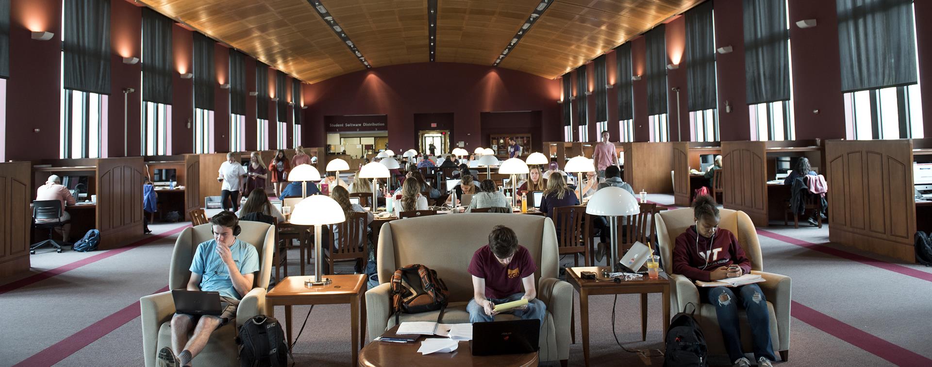 An image is students studying in the library at Virginia Tech. People sit with books and computers in front of them at tables.