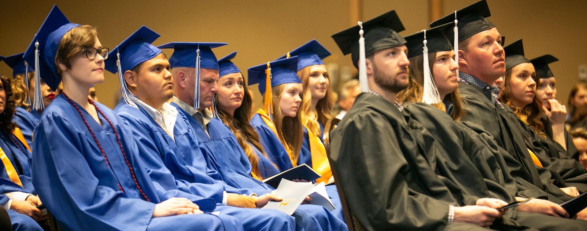 A group of students sit at graduation wearing cap and gowns.