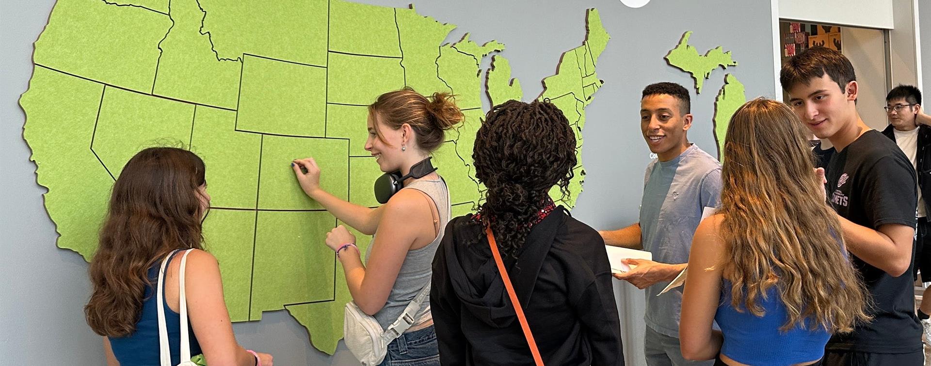 A group of six college students gathers around a large green felt map of the United States. One places a pin into Colorado.